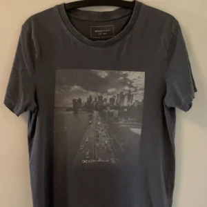 Tom Tailor t-shirt - Snygg mörkgrå t-shirt från Tom Tailor med ett stort svartvitt stadsmotiv på bröstet. Klassisk rund halsringning och korta ärmar. Perfekt för dig som gillar streetstyle och vill ha en cool vardagslook.