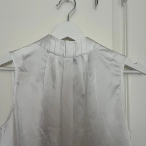 Vit ärmlös blus från STOCKH LM - Säljer en stilren vit blus från STOCKH LM i storlek S. Blusen är ärmlös med hög krage och har en mjuk, glansig finish som ger en lyxig känsla. Materialet är lent och har ett lätt fall. Perfekt för dig som gillar minimalistisk och chic stil.