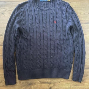 Mörkblå kabelstickad tröja Polo Ralph Lauren - Kabelstickad mörkblå tröja från Polo Ralph Lauren med klassisk rund halsringning och röd broderad logga på bröstet. Tröjan har långa ärmar och ribbade muddar för en snygg passform. Perfekt för lager-på-lager under kyliga dagar.