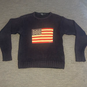 Mörkblå stickad tröja Polo Ralph Lauren - Mörkblå stickad tröja från Polo Ralph Lauren med amerikansk flagga i rött, vitt och blått på bröstet. Klassisk rund halsringning och ribbade muddar vid ärmslut och nederkant. Perfekt för dig som gillar preppy stil och vill sticka ut.