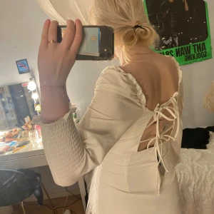 Krämvit kortklänning med snörning - Supersöt krämvit kortklänning från DK Dima med smockade trekvartsärmar, volangdetaljer vid halsringningen och snygg snörning i ryggen. Klänningen har en lätt ribbad struktur och öppen rygg som ger en romantisk vibe.