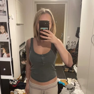 Grå ribbad linnetopp från Bershka - Säljer en grå ribbad linnetopp från Bershka med rundad hals och knappdetalj framtill. Toppen är croppad och har breda axelband, perfekt för dig som gillar enkel och stilren look. Materialet är mjukt och stretchigt bomull.