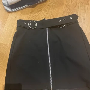 Svart kortkjol med bälte och dragkedja - Svart kortkjol med coolt bälte med silverringar och öglor i midjan. Dragkedja framtill som ger en edgy vibe. Perfekt för dig som gillar en stilren men ändå tuff look.