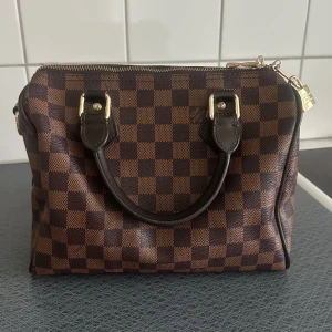 handväska brun - Snygg handväska  i klassiskt brunt Damier-mönster. Väskan har två robusta handtag, guldfärgade metalldetaljer och dragkedja med lås. Tillverkad i canvas med detaljer i skinn, perfekt för dig som gillar lyxiga accessoarer.