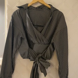 Mörkgrå skjorta med vita ränder från SHEIN - Säljer en mörkgrå skjorta från SHEIN med vita smala ränder. Skjortan har långa ärmar, krage och bred knytning i midjan som ger en snygg siluett. Materialet är mjukt och lätt, perfekt för dig som gillar trendiga detaljer och stilren look.