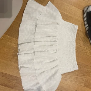 Vit volangkjol med smock - Supersöt vit kortkjol med bred smock i midjan och volanger. Kjolen har ett diskret rutmönster i tyget och är perfekt för en fräsch och somrig look. Lätt och luftig känsla, passar perfekt till sneakers eller sandaler.