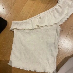 Vit oneshoulder topp med volang - Säljer en vit oneshoulder topp med volangkant längs axeln och nederkanten. Toppen är ärmlös och har en lätt skrynklig struktur i tyget som ger en cool och avslappnad vibe. Perfekt för dig som gillar att sticka ut med en unik detalj på din outfit.