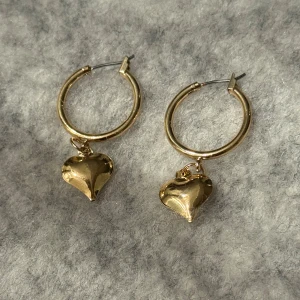 Guldiga örhängen med hjärtanhänge - Snygga guldfärgade hoops med blanka hjärtan som hänger i varje ring. Örhängena har en klassisk rund form och hjärtana ger en söt och trendig touch. Perfekta för att lyfta vilken outfit som helst.