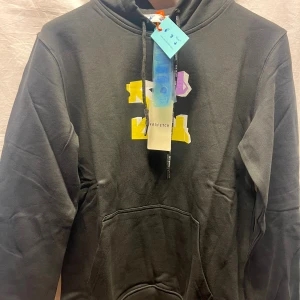 Svart hoodie från Off med tryck - Svart hoodie från Off med färgglatt tryck i gult och lila framtill och grafiskt motiv med text på ryggen. Klassisk känguruficka, huva med snörning och mjukt bomullsmaterial. Perfekt för dig som gillar streetwear vibes och vill sticka ut lite extra.