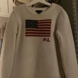 Vit stickad tröja Polo Ralph Lauren - Vit stickad tröja från Polo Ralph Lauren med amerikansk flagga och RL-broderi på bröstet. Klassisk rund halsringning och ribbade muddar. Passar 8-10 åring