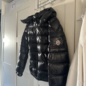 Svart Moncler maya jacka  - Säljer en svart pufferjacka från Moncler med glansig finish och huva. Jackan har dragkedja framtill, fickor med dragkedja och Moncler-logga på ärmen. Endast använd några fåtal gånger den är som i nyskick! Jag har vanligtvis storlek xs/s så skulle säga att den är där någonstans i storleken! Har även kvar prislappar och kvitto på jackan.