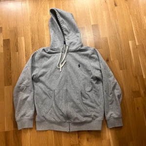 Grå hoodie från Ralph Lauren - Grå hoodie från Ralph Lauren med dragkedja och vit snörning i huvan. Klassisk broderad logga på bröstet och två stora fickor framtill. Mjuk bomullskvalitet och ribbade muddar vid ärmslut och nederkant. Perfekt för chill dagar.