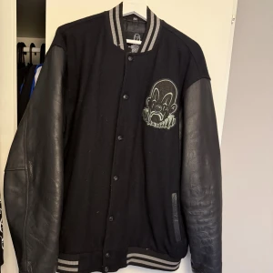 Joker brand College jacka  XXL med läderärmar - Säljer en svart bomberjacka från Joker Brand i storlek XXL. Jackan har ärmar i äkta skinn, kropp i ullblandning och gråsvarta ribbade muddar. På bröstet finns en broderad clown-logga. Klassisk collegekrage och knäppning med knappar framtill.