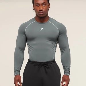 Grå baselayer Gymshark - Slimmad grå longsleeve baselayer från Gymshark med vita kontrastsömmar och logga på bröstet. Tillverkad i stretchigt syntetmaterial som sitter tight mot kroppen och passar perfekt för gym eller sport. Rund halsringning och diskret logga i nacken. Enbart testad! Original paketering medföljer! 