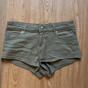 Olivgröna lågmidjade jeansshorts - Snygga olivgröna shorts i jeansmaterial med klassisk femficksdesign och knappstängning fram. Modellen är lågmidjad och har bälteshällor, perfekt för dig som gillar en enkel och clean look. Passar dig som vill ha ett par korta shorts med lågmidjade