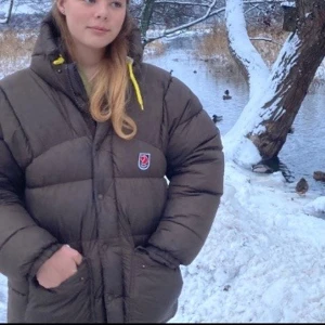 Fjällräven Jacka - Säljer en brun, super varm dunjacka från Fjällräven med huva och gula snören. Jackan har broderad logga på bröstet, stora fickor med knappar och dragkedja framtill. Perfekt för kalla vinterdagar och har en loose passform som passar lite stor på mig som brukar ha storlek S/M. Jackan är storlek XXS. Skriv gärna om du har frågor!💕 Jackan är nyligen kemtvättad och inte används sen dess!🫧