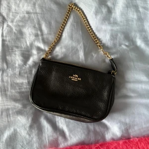 Svart axelväska från Coach  - Snygg svart axelväska från Coach i äkta skinn med guldfärgad kedja som axelrem. Väskan har ett stilrent, klassiskt utseende med Coach-logga i guld på framsidan och dragkedja upptill. Perfekt storlek för det viktigaste och ger en lyxig vibe.                  ✨Pris kan diskuteras✨