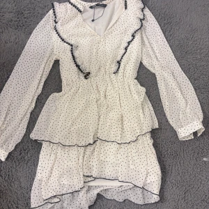 Vit prickig volangklänning från Zara - Supersöt vit kortklänning från Zara med svarta prickar, volanger och svarta kanter. Klänningen har långärmade ärmar, v-ringning och omlottkänsla framtill. Andvänd 1 gång!!