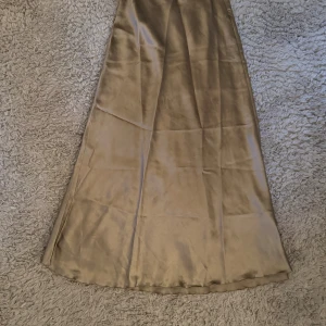 Champagnefärgad långkjol i satin - Säljer en elegant, champagnefärgad långkjol från The Black Label Collection. Kjolen har en glansig satinfinish och en mjukt utställd siluett som ger ett lyxigt intryck. Perfekt för dig som gillar stilrena och trendiga plagg.