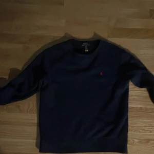 Mörkblå tröja från Polo Ralph Lauren - Snygg mörkblå tröja från Polo Ralph Lauren med klassisk rund hals och lång ärm. Tröjan har det ikoniska röda Polo-broderiet på bröstet och är gjord i mjuk bomull. Perfekt för dig som gillar stilrena och tidlösa plagg med en sportig touch.