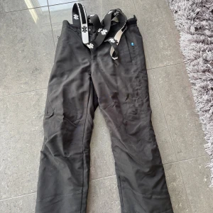 Svarta täckbyxor från Ski Industries - Svarta täckbyxor från Ski Industries med justerbara hängslen i svart och vitt. Byxorna är vindtäta och vattenavvisande, perfekta för kalla dagar. De har en enkel design med diskret logga och praktiska detaljer för skidåkning eller vinteraktiviteter.