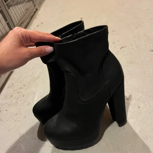 Svarta stövletter med hög klack - Säljer ett par svarta stövletter med chunky hög klack och plattform. Skorna har rund tå och dragkedja på insidan för enkel påtagning. Tillverkade i ett matt material som ger en stilren look, perfekta för att lyfta vilken outfit som helst.
