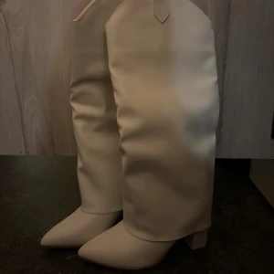 Vita höga boots med klack - Säljer ett par snygga höga boots i vit färg med spetsig tå och bred klack. Skorna är tillverkade i syntetmaterial och har en stilren, minimalistisk look som sticker ut. Perfekta för dig som vill ha ett statement-plagg till din outfit.