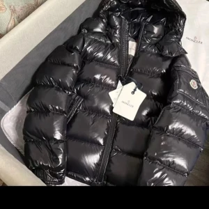 Svart pufferjacka från Moncler - Säljer en svart, glansig pufferjacka från Moncler med huva och tydlig logga på ärmen. Jackan har grov dragkedja med dubbla zippar och är riktigt fluffig och varm. Perfekt för kalla dagar och har en modern, oversized vibe.