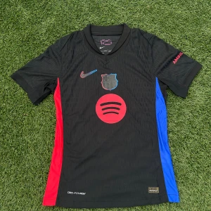 FC Barcelona svart Nike fotbollströja - Svart FC Barcelona fotbollströja från Nike med Dri-FIT ADV-teknologi. Tröjan har korta ärmar, röda och blå sidopaneler, tryckt Spotify-logga på bröstet och UNHCR-tryck på ryggen. Materialet är lätt och andas, perfekt för match eller träning.