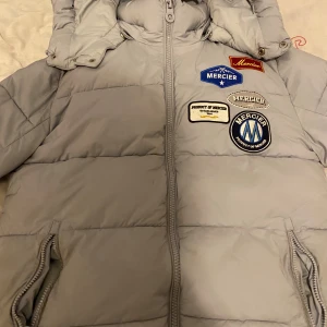 Ljusgrå pufferjacka från Mercier - Snygg ljusgrå pufferjacka från Mercier med flera broderade patches på bröstet. Jackan har huva, dragkedja framtill och två fickor med dragkedja. Perfekt för kalla dagar och ger en cool streetstyle-vibe.