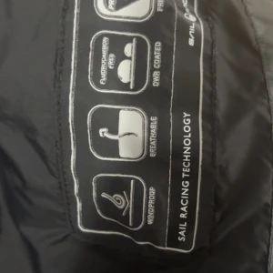 Svart vindjacka från Sail Racing - Svart vindjacka från Sail Racing med huva och minimalistisk design. Jackan är tillverkad i lätt och slitstark polyester, är vindtät och andas. Perfekt för blåsiga dagar och har Sail Racing-logga på huvan. Snygg och sportig look.