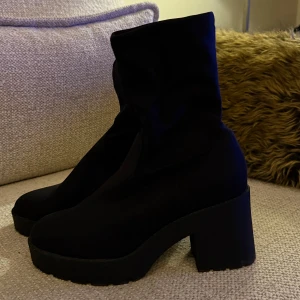 Svarta boots med hög klack - Säljer ett par svarta boots med stretchig skaft och chunky hög klack. Skorna har rund tå och platt sula framtill, vilket ger en cool och modern siluett. Materialet är mjukt och matt, perfekt för dig som gillar minimalistisk stil.