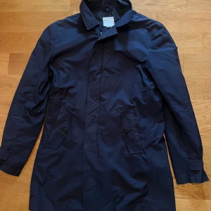 Marinblå rock från J.Lindeberg - Snygg marinblå rock från J.Lindeberg med klassisk krage och två fickor framtill med knappar. Jackan har diskret logodetalj vid kragen och stängs med både dragkedja och knappar. Perfekt för dig som gillar stilren och tidlös design.