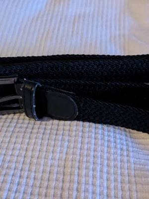 Svart flätad bälte i textil och skinn - Säljer ett svart flätat bälte med detaljer i skinn och metallspänne. Bältet är i textilmaterial med en snygg struktur och har en klassisk look som passar till jeans eller chinos. Längd 90 cm.