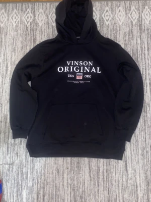 Svart hoodie från Vinson Original - Svart hoodie från Vinson Original med stor vit text och amerikanskinspirerad logga på bröstet. Klassisk känguruficka framtill och huva. Tillverkad i mjukt material, perfekt för en avslappnad och cool stil.