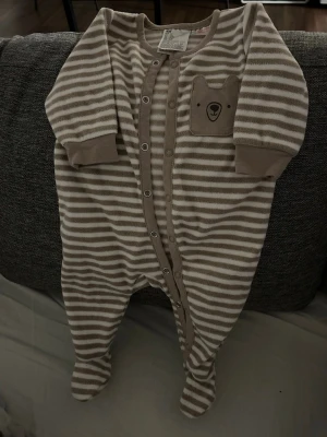 Bebiskläder Randig beige och vit babyoverall H&M - Mysig babyoverall från H&M i storlek 62. Overallen är beige och vit med horisontella ränder, har tryckknappar framtill och en söt björn-detalj på bröstet. Tillverkad i mjuk polyester och har inbyggda fötter.