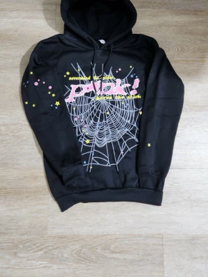 Svart hoodie med spindelnätsmönster - Säljer en svart hoodie med stor huva och tryck framtill. Motivet består av ett vitt spindelnät, rosa text och små gula och rosa stjärnor. Texten säger 'never mind the spider, here’s the web!'. Perfekt för dig som gillar coola och unika prints.