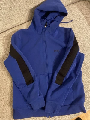 Blå hoodie från BOSS med svarta detaljer - Säljer en blå hoodie från BOSS med svarta paneler på ärmarna och dragkedja framtill. Tröjan har huva med snörning och matchande blå byxor med svarta detaljer. Perfekt för chill eller streetwear. Materialet är mjukt och känns som bomull eller polyester. Nypris 3400 kr pris kan diskuteras