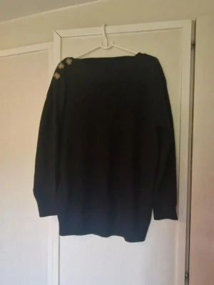 Svart stickad tröja med dekorativa knappar - Mysig svart stickad tröja med tre dekorativa knappar på höger axel. Tröjan har en rak passform, långa ärmar och bred halsringning. Perfekt för lager på lager och enkel att styla till olika outfits. Utöver att den är lite nopprig så hittar jag inga skavanker.