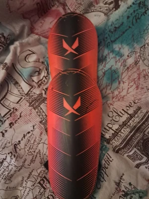 Röda och svarta benskydd från X - Säljer ett par sportiga benskydd i rött och svart med coolt grafiskt mönster och X-logga framtill. De har en böjd form som sitter skönt på benet och är tillverkade i ett tåligt plastmaterial, perfekta för fotboll eller andra sporter där skydd behövs.