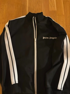 Palm Angels svart track jacket - Säljer en svart track jacket från Palm Angels med vita ränder längs ärmarna och vit dragkedja framtill. Klassisk hög krage och logga på bröstet. Perfekt för en sportig och trendig look.