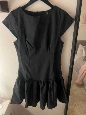 Svart kort volangklänning - Stilren svart kort klänning med volang nedtill och rund halsringning. Klänningen har korta ärmar och en figurnära passform upptill. Tillverkad i ett slätt tyg som ger en elegant känsla. Perfekt för dig som gillar minimalistisk och chic stil.