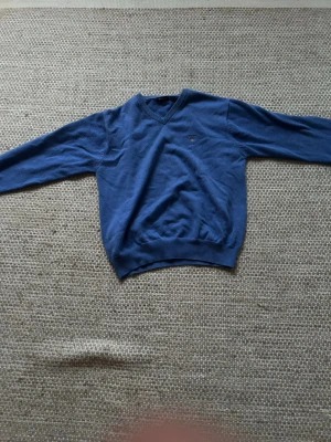 Blå stickad tröja med V-ringning från Gant - En klassisk blå stickad tröja med V-ringad hals och långa ärmar. Tröjan har ribbade muddar vid ärmslut och nederkant samt en liten broderad Gant logga på bröstet. Perfekt för lager-på-lager och enkel att matcha med jeans eller chinos.
