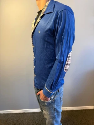 Burberry skjorta - Snygg blå skjorta från Burberry med klassiska rutiga detaljer på krage, knappslå och armbågslappar. Storlek M. Modellen är 184cm.