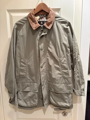 Grön rock från Barbour med manchesterkrage - Klassisk grön rock från Barbour med beige manchesterkrage och rutigt innerfoder. Rocken har stora fickor framtill, knappar och dragkedja. Perfekt för dig som gillar tidlös brittisk stil och vill ha en rock med snygga detaljer.