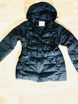 Svart dunjacka från Moncler med huva - Snygg svart dunjacka från Moncler med quiltad design och praktisk huva. Jackan har dragkedja framtill, flera fickor och en klassisk Moncler-logga på insidan. Perfekt för kalla dagar och ger en stilren look.