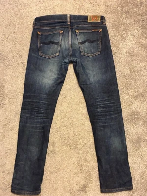 Nudie jeans vintage - Säljer ett par mörkblå jeans från Nudie med klassiska kontrastsömmar och slitningar framtill. Jeansen har fem fickor och är tillverkade i stretchig bomullsdenim för en bekväm passform.