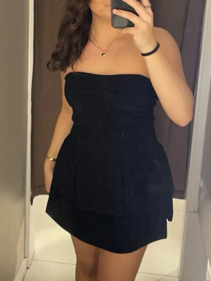 Svart axelbandslös klänning från Missäy - Säljer en svart axelbandslös kort klänning med volangdetaljer från Missäy. Klänningen har en figurnära passform upptill och en lätt utsvängd kjol. Har vadderingen vid bysten. Den är svart med små glittriga detaljer och är onesize men i referens till andra klänningar sitter den som en XS/S (36) modell. 