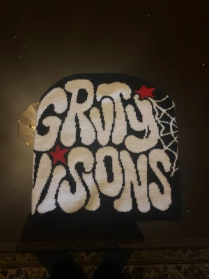 Svart mössa Groty Visions spindelnät - Säljer en svart mössa med texten 'Groty Visions' i vit, stora bokstäver och röda stjärnor. Ena sidan har ett coolt spindelnätsmönster. Perfekt för dig som gillar streetwear och unika accessoarer.
