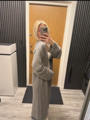 Grå lång kappa i ullmix - Snygg och stilren grå kappa i lång modell med avslappnad passform. Kappan har långa ärmar, öppen framtill och är tillverkad i mjuk ullmix som känns både varm och bekväm. Perfekt för dig som gillar en minimalistisk look. Mohair och wool